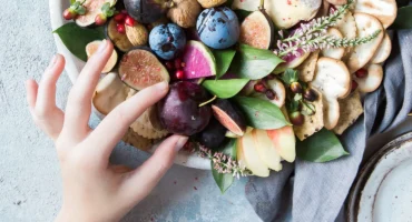 Teller mit Früchten, Nüssen und Käse – eine ausgewogene Mahlzeit, die Kohlenhydrate und gesunde Fette kombiniert, mit einer Hand, die eine Zwetschge nimmt.