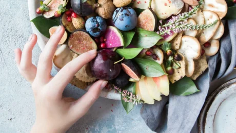 Teller mit Früchten, Nüssen und Käse – eine ausgewogene Mahlzeit, die Kohlenhydrate und gesunde Fette kombiniert, mit einer Hand, die eine Zwetschge nimmt.